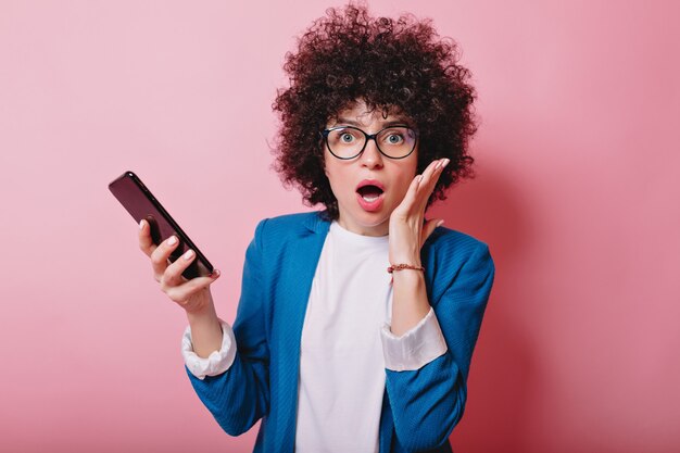 La donna sorpresa con la giacca blu vestita capelli corti ondulati tiene lo smartphone con la bocca aperta sul colore rosa