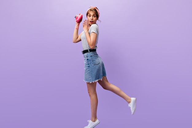 La donna sorpresa in gonna di jeans tiene la macchina fotografica rosa. Bella ragazza con fascia estiva in camicia grigia e scarpe da ginnastica bianche in posa.