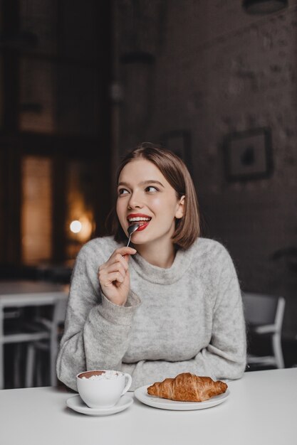 La donna sorridente con il rossetto rosso sta leccando il cucchiaino. Ragazza in abito di cashmere godendo di croissant.