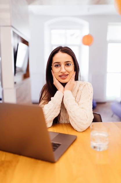 La donna sorridente con le parentesi graffe con i capelli neri di Londra sta lavorando al suo computer portatile e sta sedendosi a casa alla tavola di legno