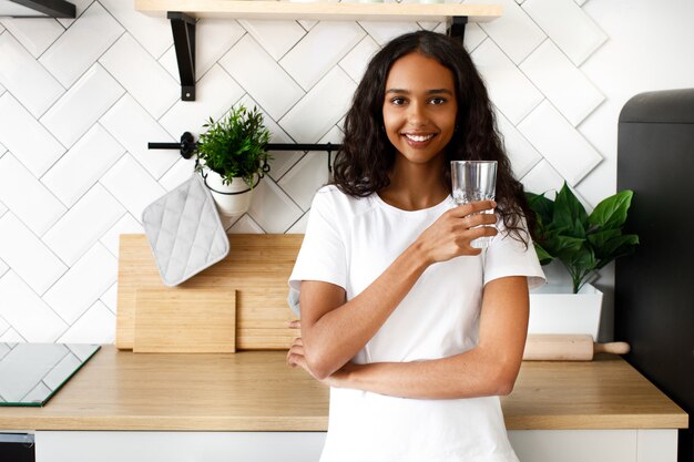 La donna sorridente del mulatto sta tenendo il vetro con acqua vicino allo scrittorio della cucina sulla cucina bianca moderna
