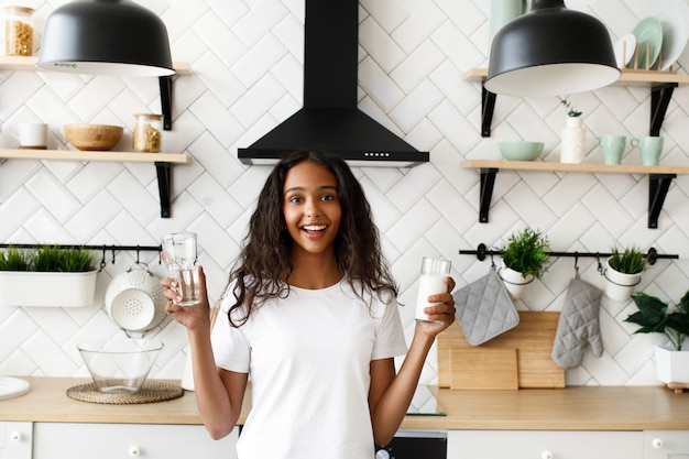 La donna sorridente del mulatto sta tenendo il vetro vuoto e il vetro con latte vicino allo scrittorio della cucina sulla cucina bianca moderna vestita in maglietta bianca