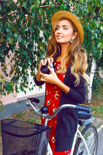 La donna sorridente felice che si diverte e sorprende le emozioni giocose, cammina da solo con la bici retrò alla moda nel parco cittadino, indossa un maglione caldo vestito rosso e un cappello di paglia vintage, ha lunghi capelli biondi arricciati.