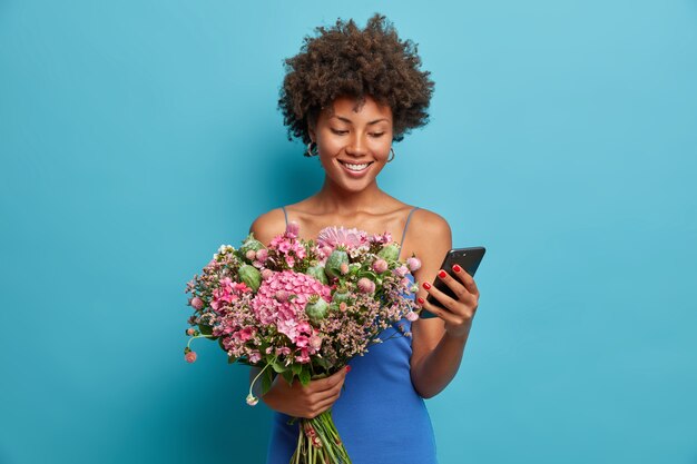 La donna sorridente felice guarda il display dello smartphone sorride ampiamente, tiene il telefono cellulare e un mazzo di fiori