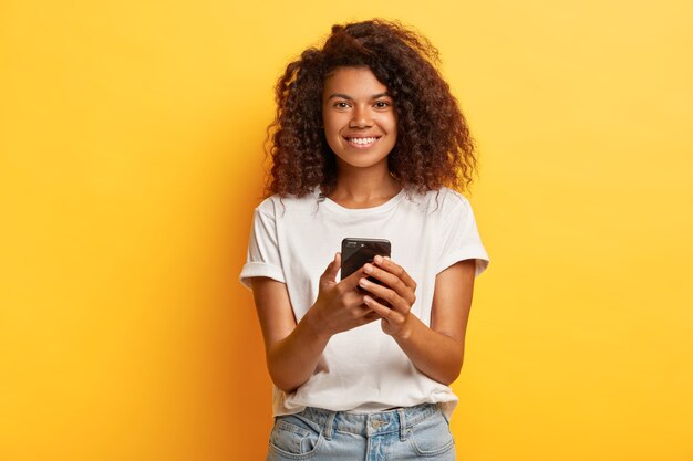 La donna sorridente felice tiene il telefono cellulare, il messaggio di testo sul cellulare, naviga in Internet, ha l'acconciatura riccia e folta, vestita con jeans e maglietta casual bianca