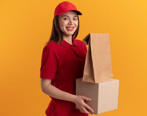 La donna sorridente graziosa delle consegne in uniforme tiene il pacchetto di carta su una scatola di cartone isolata sulla parete arancione con lo spazio della copia