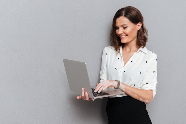 La donna sorridente in affari copre la posa vicino alla parete e per mezzo del computer portatile su gray