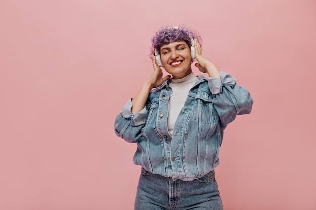 La donna sorridente in giacca di jeans blu ascolta musica con gli occhi chiusi. Donna in cuffie bianche in posa sul rosa.