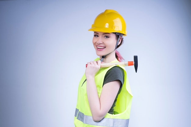 La donna sorridente in un casco giallo sta con un martello sulla sua spalla