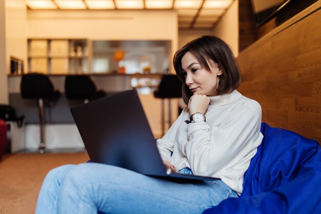 La donna sorridente sta sedendosi nella sedia della borsa blu che lavora al computer portatile brillante che parla sul telefono