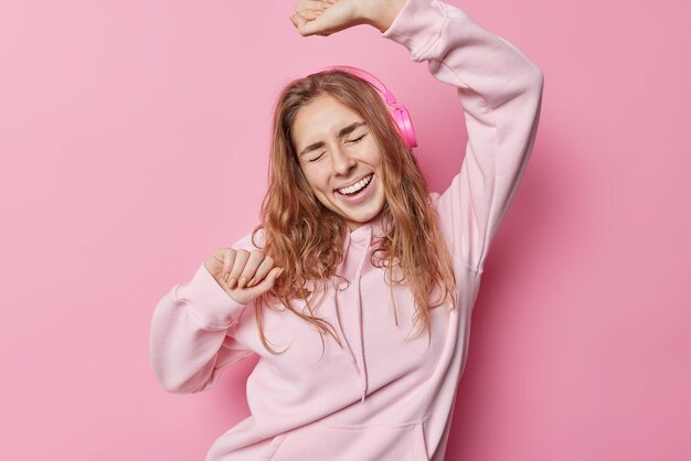 La donna spensierata positiva con i capelli lunghi vestita con felpa con cappuccio indossa le cuffie stereo balla a ritmo di musica isolata su sfondo rosa. L'adolescente felice ama le pose preferite della playlist al chiuso