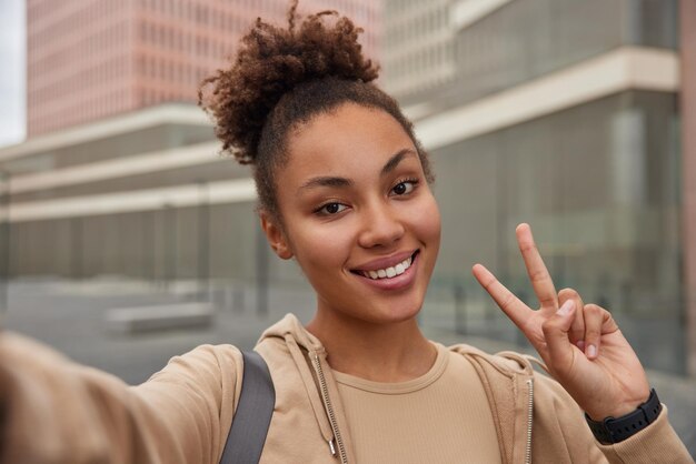 La donna spensierata positiva con i capelli ricci vestita con felpa con cappuccio sorride felicemente fa il gesto di pace si fa selfie in un luogo urbano essendo di buon umore dopo l'allenamento sportivo Emozioni della gente e stile di vita sportivo