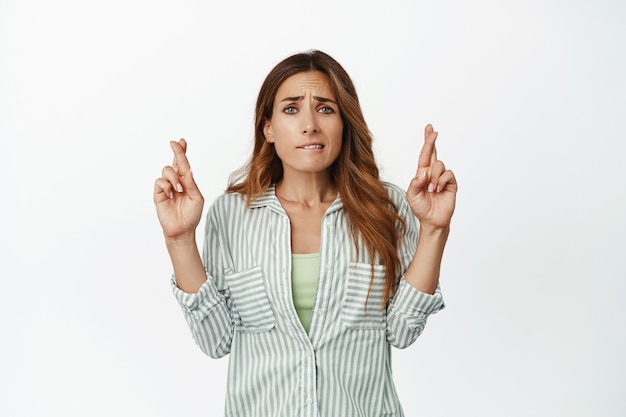 La donna speranzosa preoccupata incrocia le dita, mordendosi il labbro nervosamente, aspettando, pregando per buona fortuna, anticipando notizie positive, in piedi contro il muro bianco