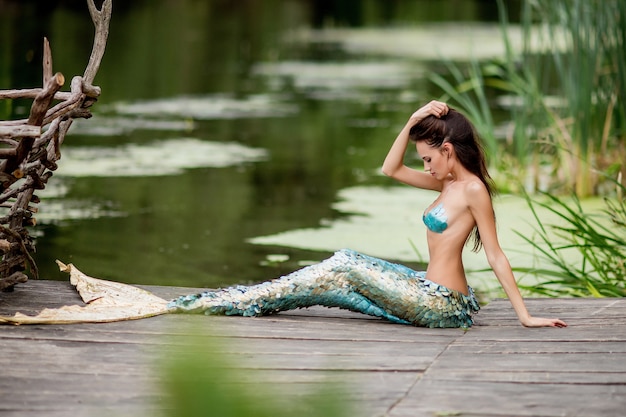 La donna splendida con i capelli lunghi e vestita come una sirena si siede sul ponte sull'acqua