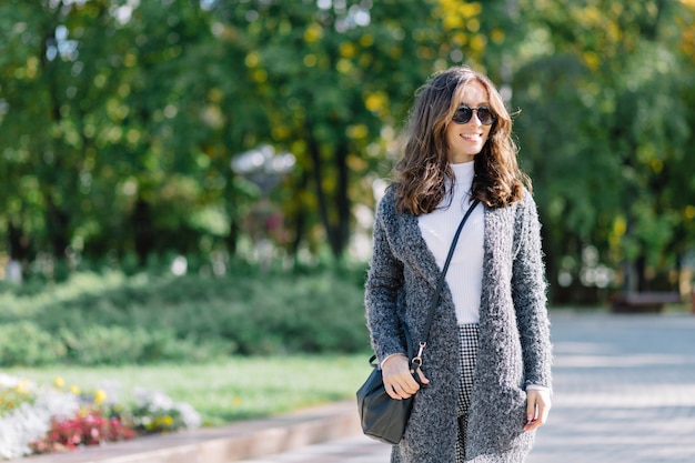 La donna sta camminando per strada con un grande sorriso. Ha i capelli corti e scuri e meravigliosi grandi occhi azzurri. È vestita con un pullover grigio e una camicia bianca.