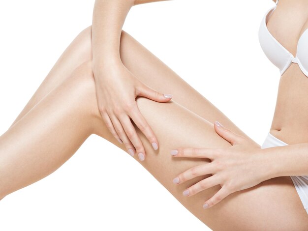 La donna stringe la pelle della cellulite sulle gambe sul muro bianco