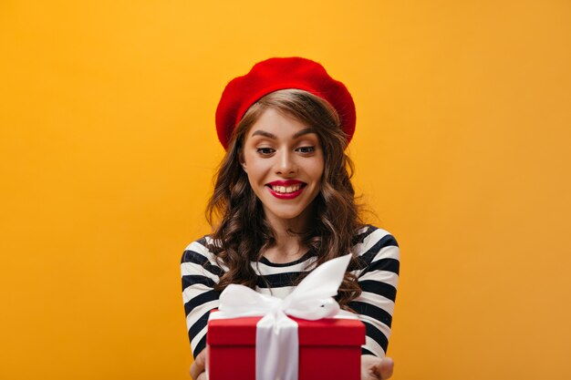 La donna stupita in berretto rosso e camicia a righe tiene il contenitore di regalo. Affascinante signora con acconciatura ondulata in cappello luminoso e camicetta moderna in posa.