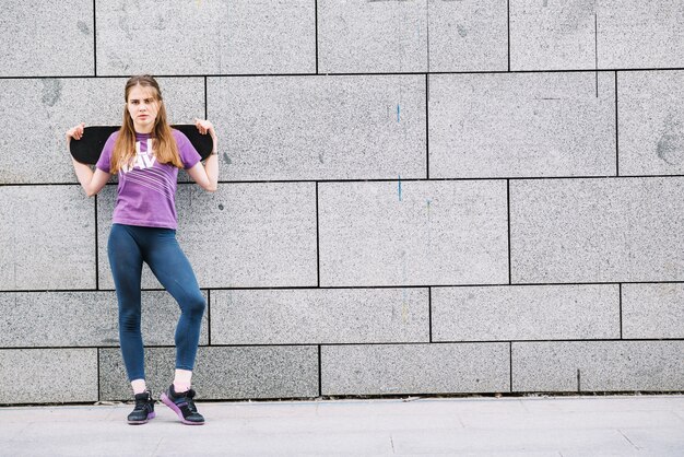 La donna tiene uno skateboard in piedi davanti ad una parete grigia