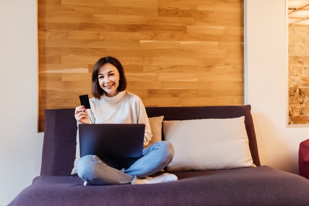 La donna vestita abbastanza casuale sta lavorando al computer portatile che si siede sul letto scuro davanti alla parete di legno a casa