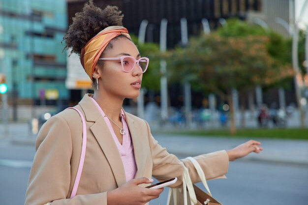 la donna vestita con abiti eleganti fa l'autostop su strada in una strada vuota indossa occhiali da sole rosa alla moda, fascia e giacca ferma le pose delle auto