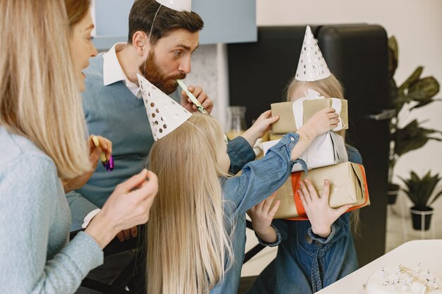 La famiglia e le due figlie festeggiano il compleanno in cucina. La gente indossa un cappello da festa. La ragazza tiene una scatola con i regali.