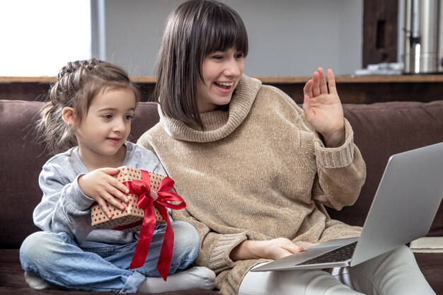 La famiglia felice che esamina lo schermo del laptop effettua la videochiamata a distanza. Madre sorridente e bambina con confezione regalo a parlare con la webcam.