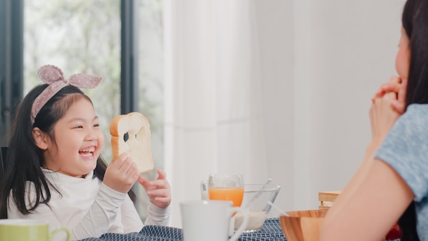 La famiglia giapponese asiatica fa colazione a casa. La figlia asiatica seleziona e gioca il sorriso di risata del pane con i genitori mentre mangia il cereale e il latte dei fiocchi di mais in ciotola sulla tavola in cucina moderna di mattina.