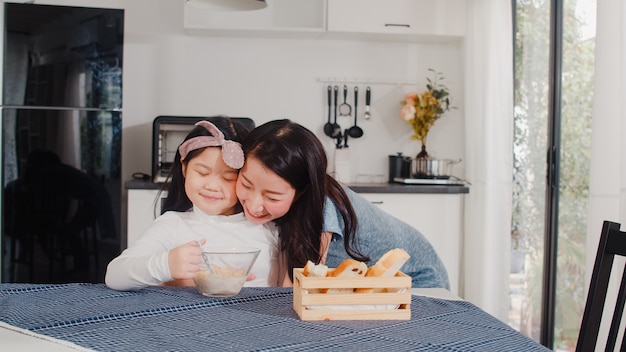 La famiglia giapponese asiatica fa colazione a casa. La mamma e la figlia asiatiche che si sentono felici parlano insieme mentre mangiano il pane, il cereale dei fiocchi di mais e il latte in ciotola sulla tavola in cucina moderna a casa nella mattina.