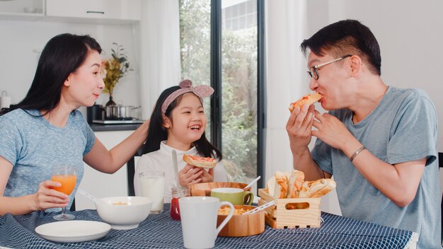 La famiglia giapponese asiatica fa colazione a casa. La mamma, il papà e la figlia asiatici che si sentono felici parlano insieme mentre mangiano il pane, i cereali dei fiocchi di mais e il latte in ciotola sulla tavola nella cucina di mattina.