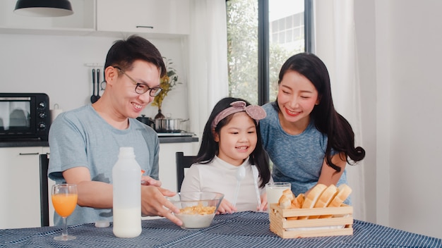 La famiglia giapponese asiatica fa colazione a casa. La mamma, il papà e la figlia asiatici che si sentono felici parlano insieme mentre mangiano il pane, i cereali dei fiocchi di mais e il latte in ciotola sulla tavola nella cucina di mattina.