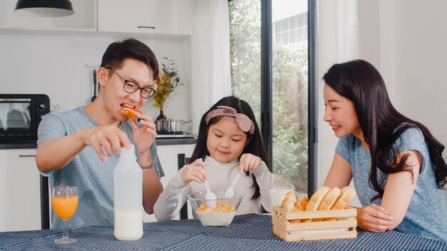 La famiglia giapponese asiatica fa colazione a casa. La mamma, il papà e la figlia asiatici che si sentono felici parlano insieme mentre mangiano il pane, i cereali dei fiocchi di mais e il latte in ciotola sulla tavola nella cucina di mattina.