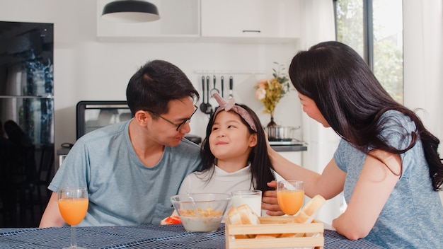 La famiglia giapponese asiatica fa colazione a casa. La mamma, il papà e la figlia asiatici che si sentono felici parlano insieme mentre mangiano il pane, i cereali dei fiocchi di mais e il latte in ciotola sulla tavola nella cucina di mattina.