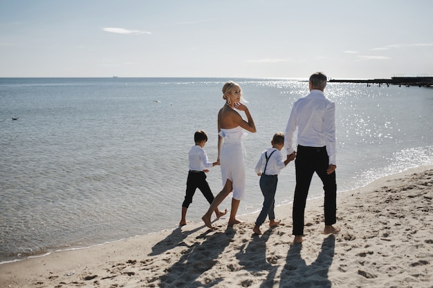 La famiglia in abiti di lusso sta camminando a piedi nudi sulla spiaggia di sabbia in una calda giornata di sole