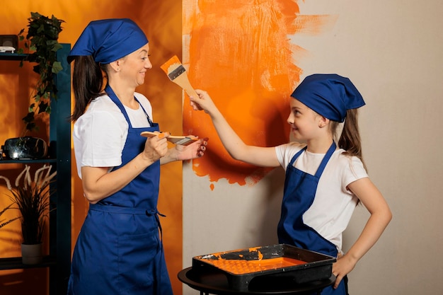 La famiglia si diverte con la vernice arancione, usando il colore per rinnovare insieme le pareti degli appartamenti. Donna con bambino che ride e dipinge la stanza della casa con gli strumenti del pennello e il lavoro interno fai da te.