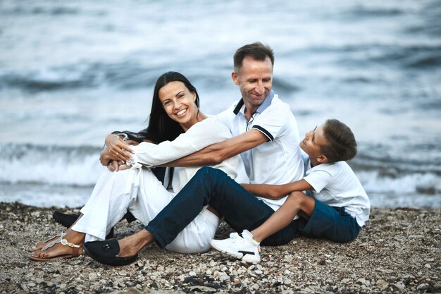 La famiglia sorrisa è seduta sulla spiaggia rocciosa vicino al mare in tempesta e abbraccia, genitori e figlio