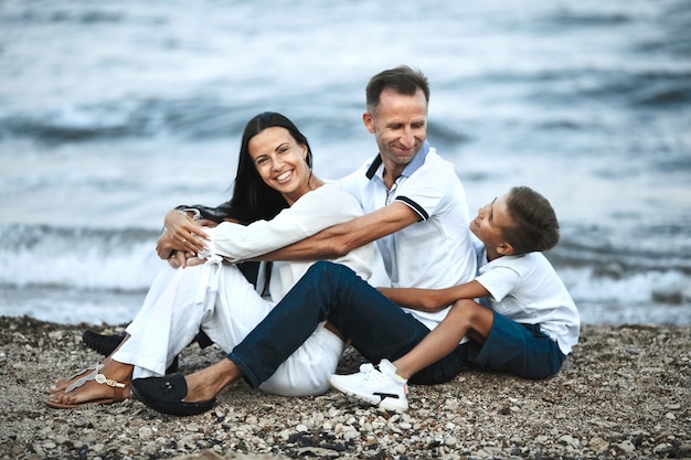 La famiglia sorrisa è seduta sulla spiaggia rocciosa vicino al mare in tempesta e abbraccia, genitori e figlio