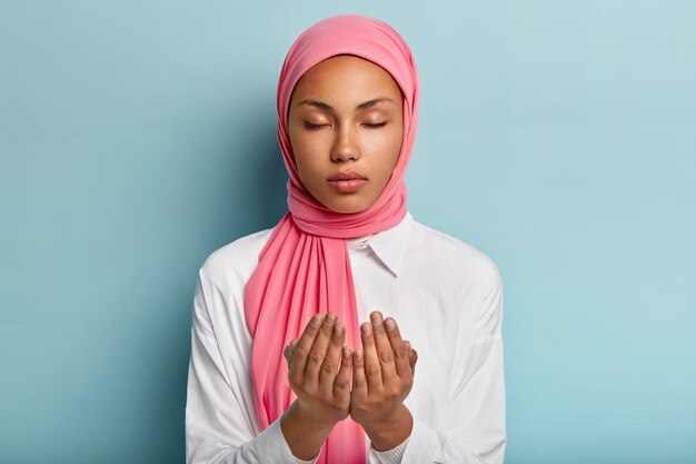 La fedele donna araba dalla pelle scura tiene le mani nel gesto di preghiera, chiede ad Allah una buona salute, crede nel benessere ha la testa velata, indossa una camicia bianca tiene gli occhi chiusi gode di un'atmosfera tranquilla