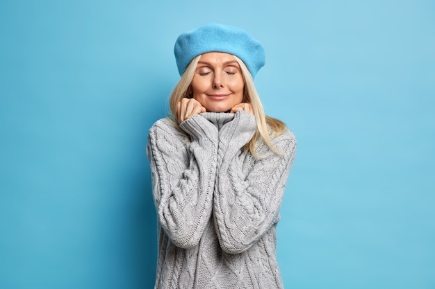 La felice donna adulta indossa un comodo maglione lavorato a maglia tiene le mani sul colletto con gli occhi chiusi e gode di un momento di pace.