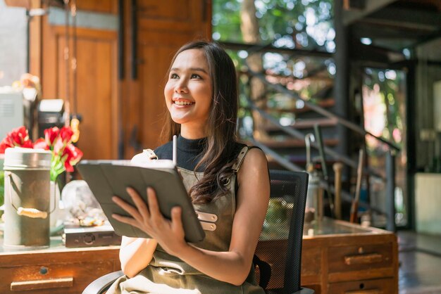 La felicità del progettista creativo adulto femminile asiatico pensa al lavoro e alla ricerca e prendi nota di idee interessanti tramite il dispositivo tabletdonna asiatica che lavora casualmente si rilassa nel suo studio di casa con giardino finestra