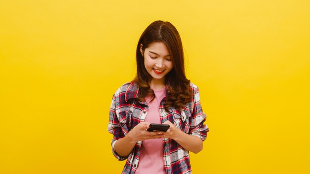 La femmina asiatica adorabile sorridente che per mezzo del telefono con l'espressione positiva, sorride ampiamente, si è vestita in abbigliamento casual ed esamina la macchina fotografica sopra la parete gialla. La donna felice adorabile felice si rallegra del successo.