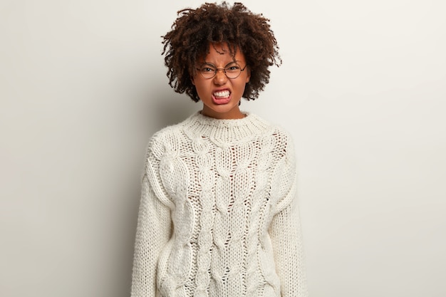 La femmina dalla pelle scura irritata fa un sorrisetto, ha uno sguardo infastidito, aggrotta le sopracciglia e stringe i denti, ha i capelli ricci, vestita con lunghi abiti invernali, isolata su un muro bianco, si sente stressata e arrabbiata