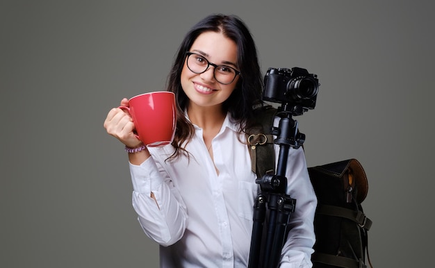La femmina del viaggiatore tiene una macchina fotografica digitale e una tazza di caffè rossa su sfondo grigio.