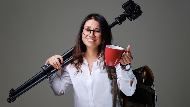 La femmina del viaggiatore tiene una macchina fotografica digitale e una tazza di caffè rossa su sfondo grigio.