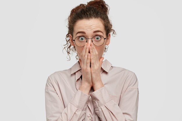 La femmina spaventata e lentigginosa guarda con gli occhi fuoriusciti, copre la bocca con i palmi delle mani, ha i capelli scuri, sente notizie terribili, indossa una camicia oversize alla moda, isolata su un muro bianco. Omg, è terribile!