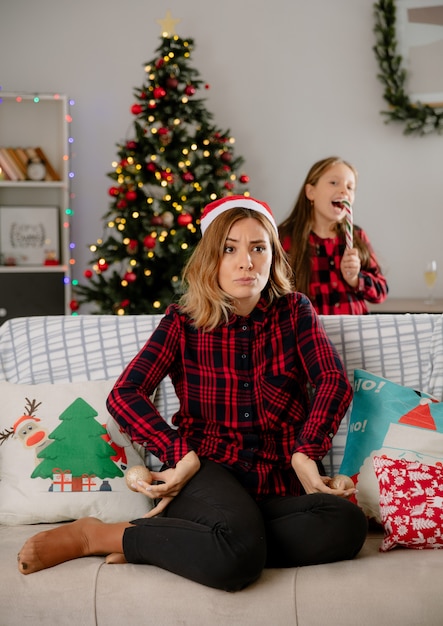 La figlia allegra che mangia il bastoncino di zucchero e la madre arrabbiata tiene gli ornamenti della sfera di vetro che si siedono sul divano e si godono il periodo natalizio a casa