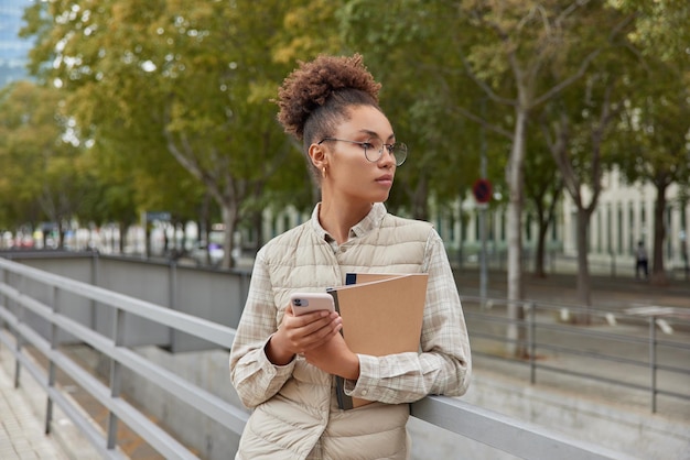 La foto all'aperto di una donna dai capelli ricci premurosa usa il telefono cellulare per chattare online tiene i quaderni in posa in città contro il parco indossa occhiali rotondi e il gilet ha un'espressione pensierosa