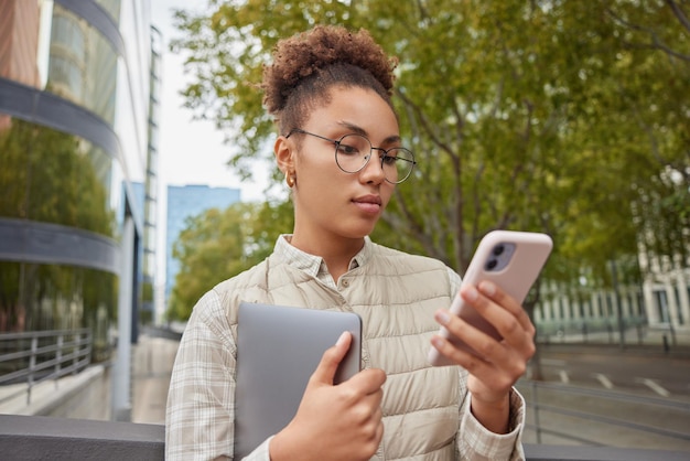 La foto all'aperto di una donna seria usa gadget moderni cammina per strada usa il telefono cellulare porta il tavolo digitale indossa una maglia e occhiali rotondi invia sms cerca di trovare un percorso in un luogo sconosciuto