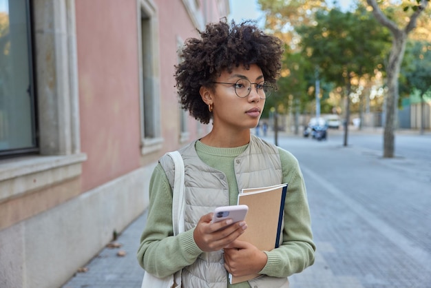 La foto all'aperto di una giovane donna riccia premurosa si gode il tempo libero in una strada cittadina usa lo smartphone tiene i taccuini focalizzati a distanza vestito casualmente L'influencer femminile condivide i contenuti nel blog