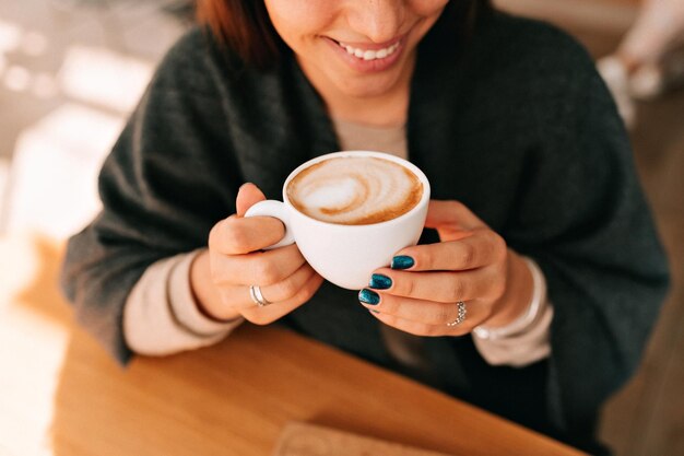 La foto dall'alto di una donna adorabile sorridente con un sorriso meraviglioso e una manicure scura tiene in mano il caffè e si gode il riposo nella caffetteria