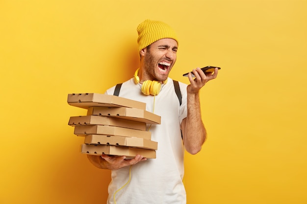 La foto del corriere infastidito dell'uomo della pizza urla con rabbia allo smartphone, ha una conversazione fastidiosa con il cliente, tiene una pila di scatole di cartone, indossa un cappello e una maglietta bianca, isolato sul muro giallo